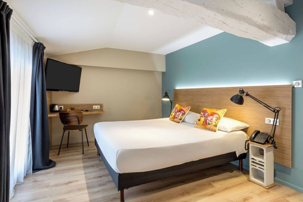 A hotel room with a bed, bedside table, lamp, and a desk with a chair.