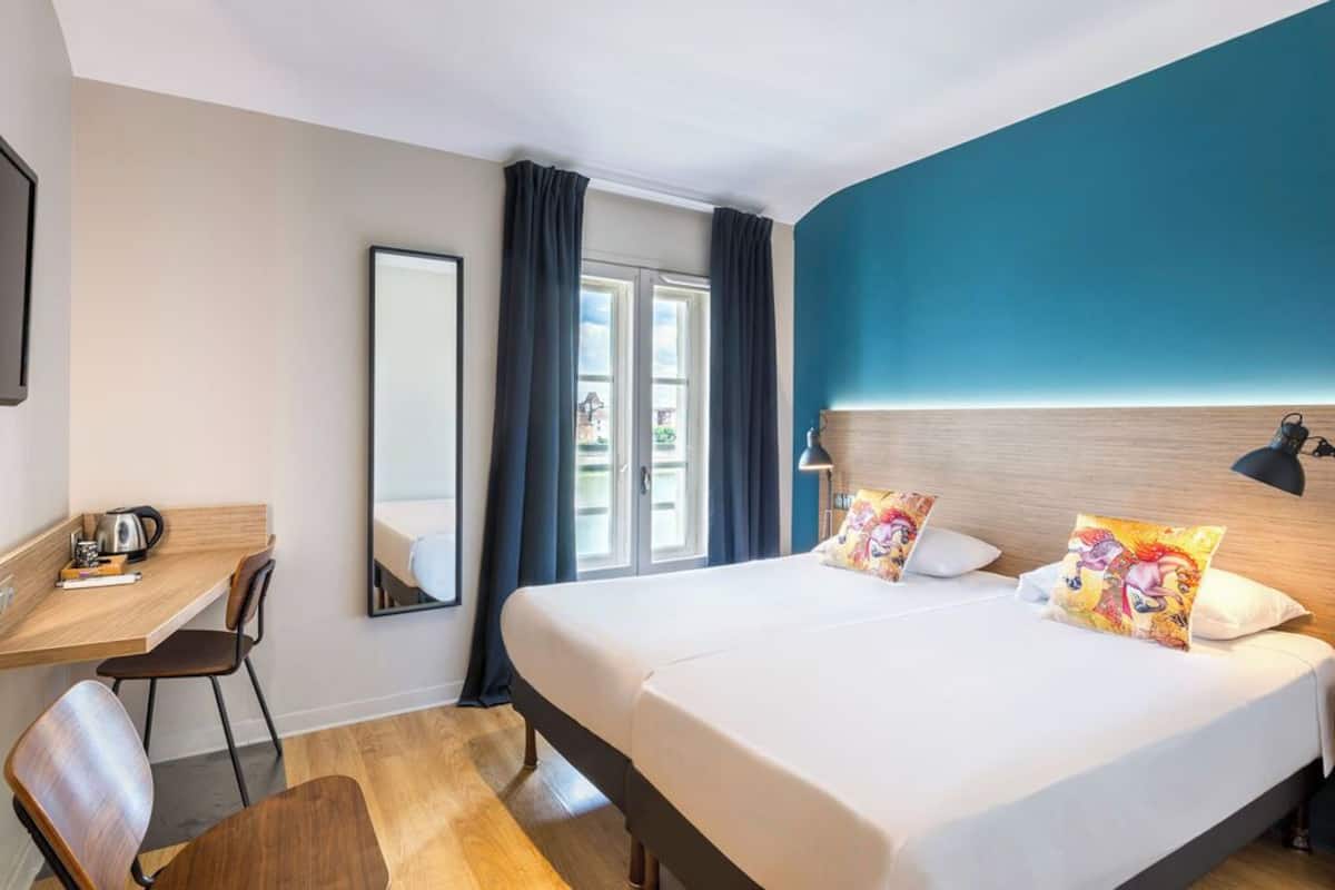 A hotel room with a bed, a desk, a chair, a mirror, and a window with curtains.