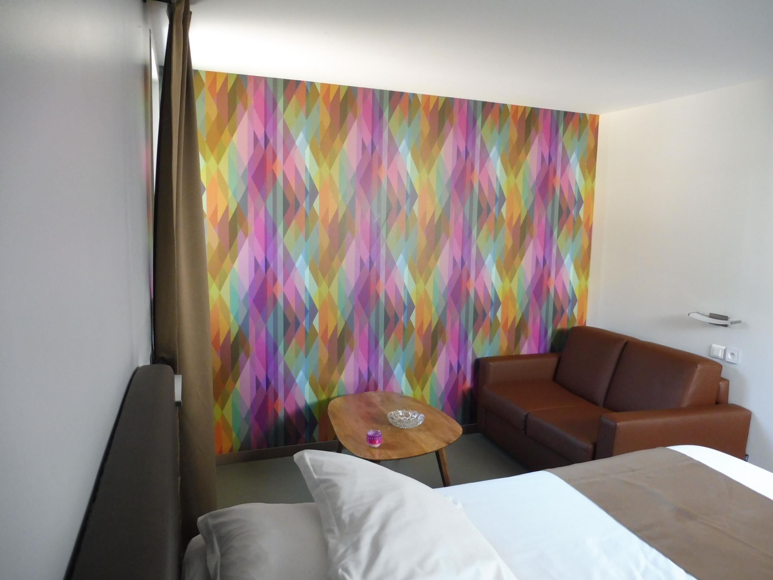 Een hotelkamer met een bed, een bruine leren bank, een klein houten tafeltje en een kleurrijk geometrisch wandontwerp.