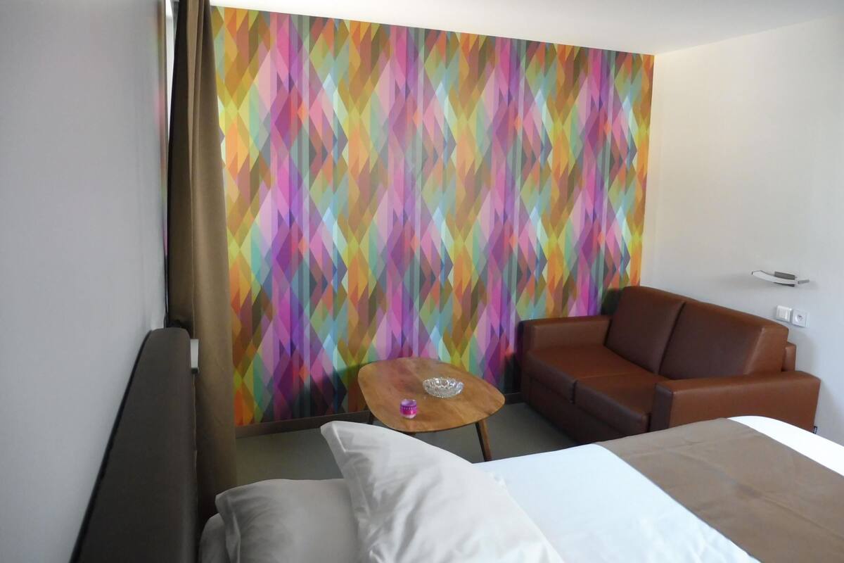 A hotel room with a bed, a brown leather sofa, a small wooden table, and a colorful geometric wall design.