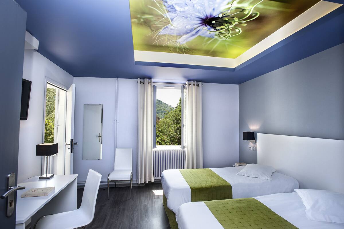 A modern hotel room with two beds, a desk, a chair, a window with curtains, and a view of greenery outside.