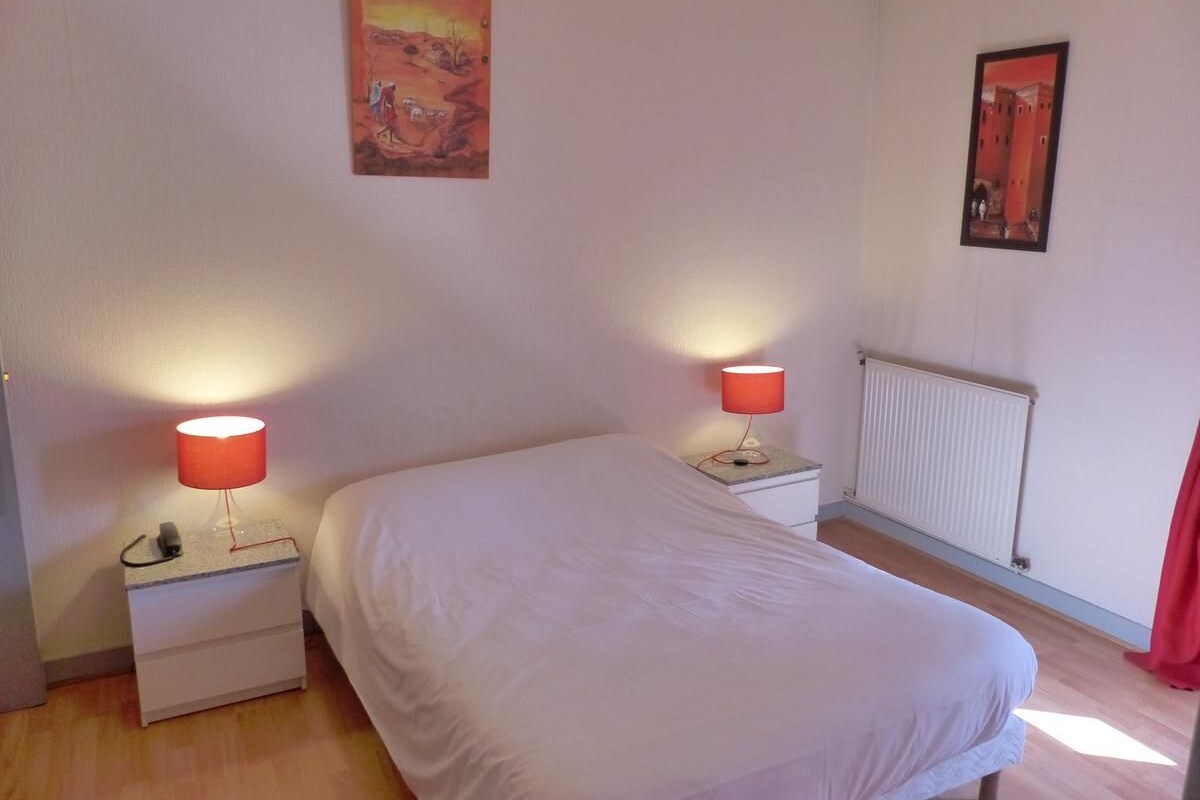A bedroom with a bed, two bedside tables with lamps, a radiator, and two paintings on the walls.