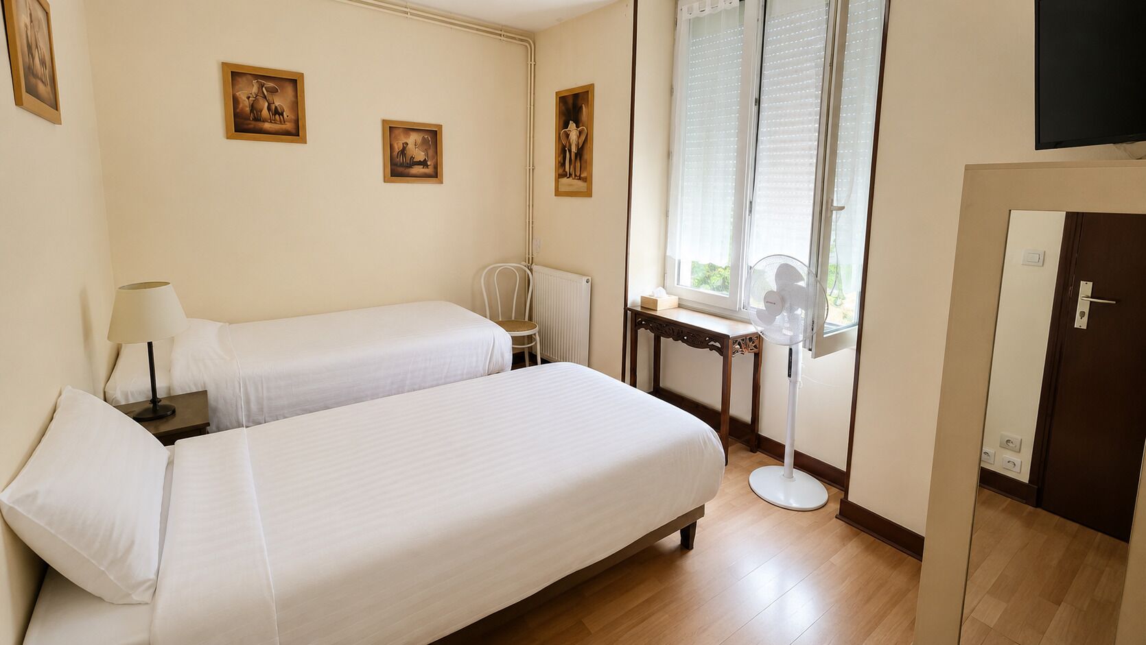 A hotel room with two beds, a bedside table with a lamp, a chair, a window with blinds, a standing fan, and a door.
