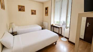 A hotel room with two beds, a bedside table with a lamp, a chair, a window with blinds, a standing fan, and a door.