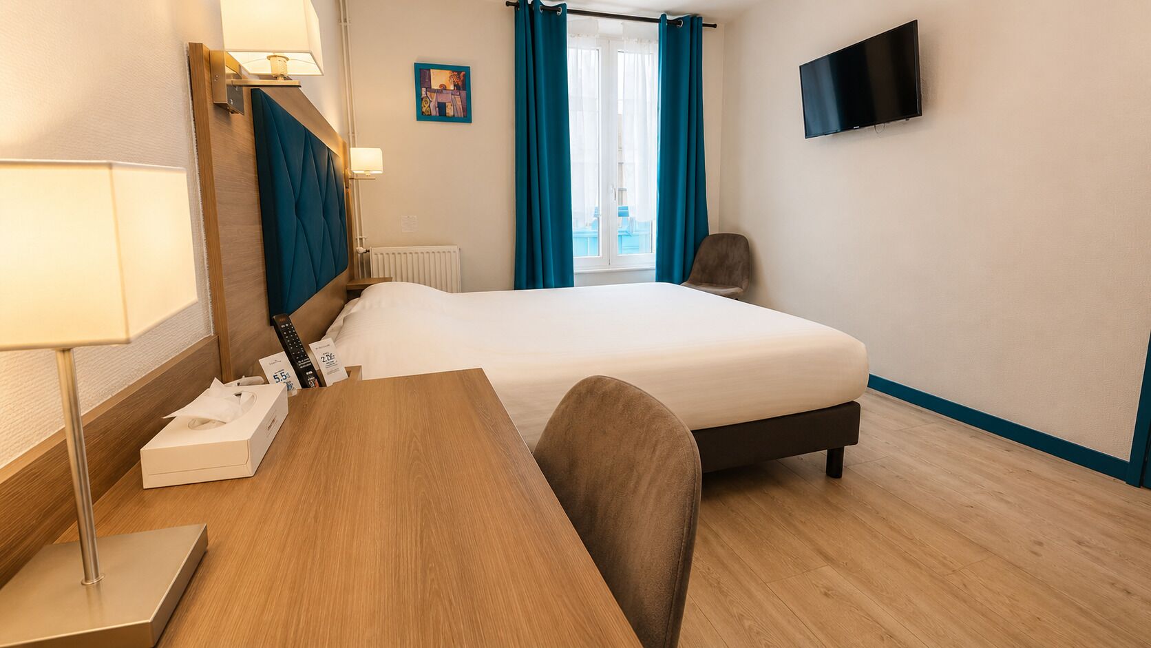 Ein Hotelzimmer mit einem Bett, einem Schreibtisch mit Lampe, einem Sessel, einem Fernseher und einem Fenster mit Vorhängen.