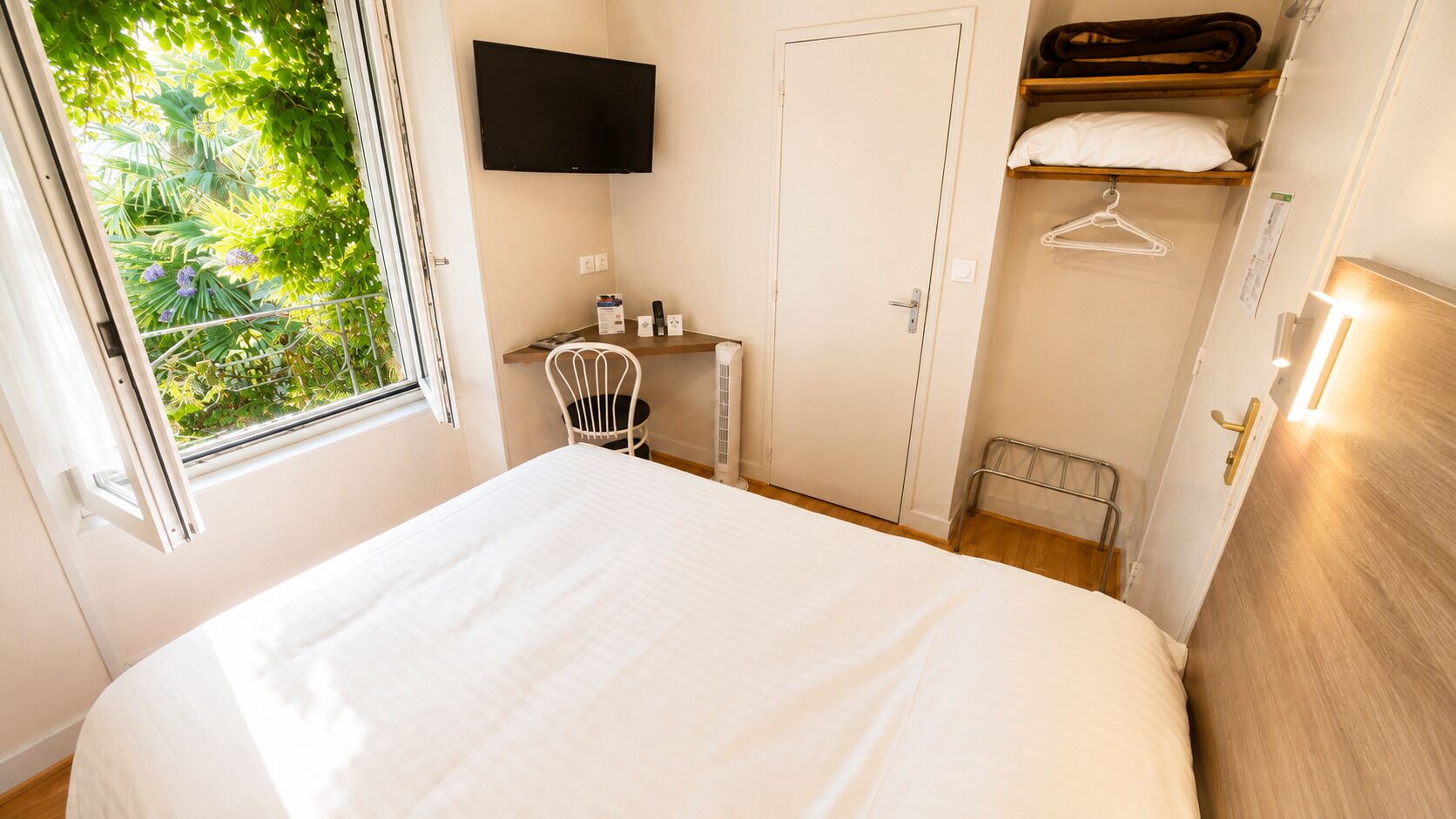 A bedroom with a bed, a chair, a TV, a wardrobe, and a window with a view of greenery.
