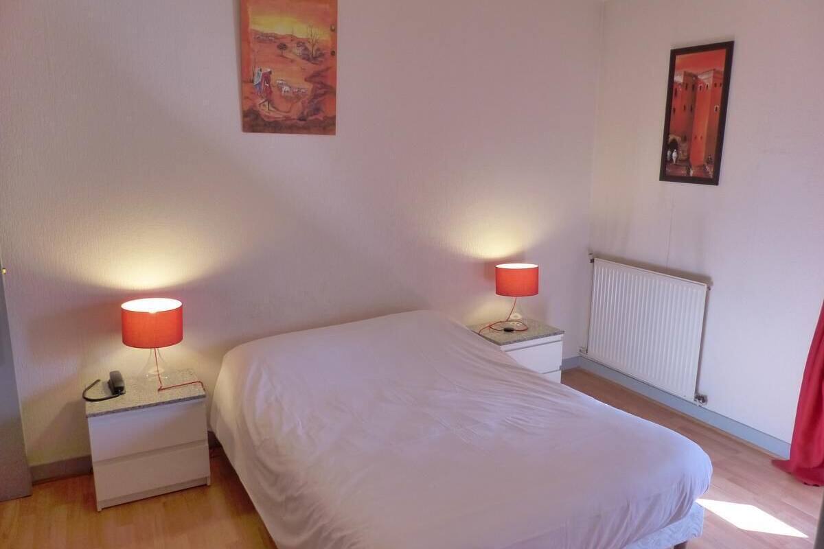 Un dormitorio con una cama, dos mesitas de noche con lámparas, un radiador y dos cuadros en las paredes.