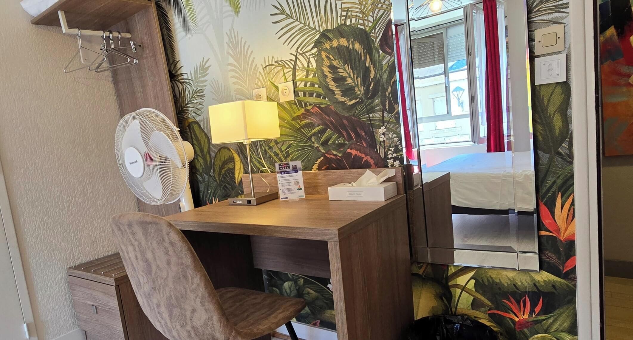 A hotel room with a wooden desk, a chair, a fan, a lamp, and a bed with a tropical wallpaper.