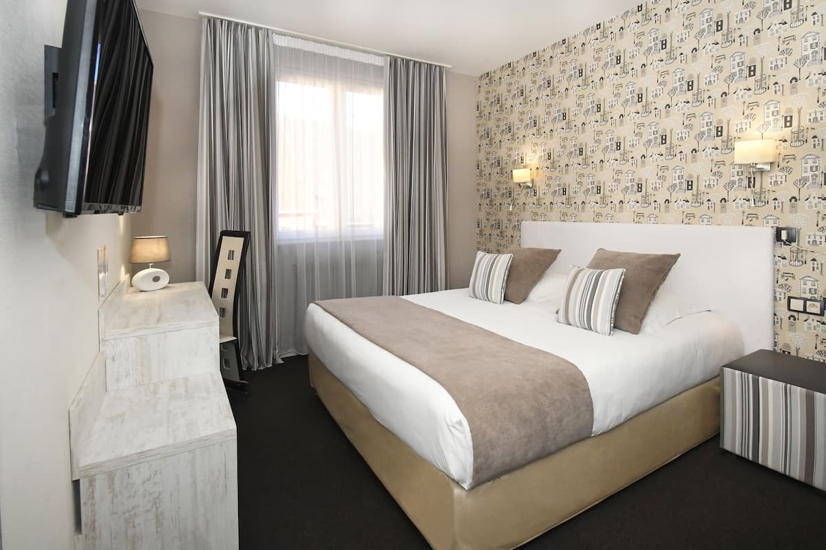 A hotel room with a large bed, a TV mounted on the wall, a desk with a lamp, and a window with curtains.