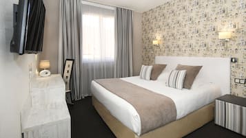 A hotel room with a large bed, a TV mounted on the wall, a desk with a lamp, and a window with curtains.