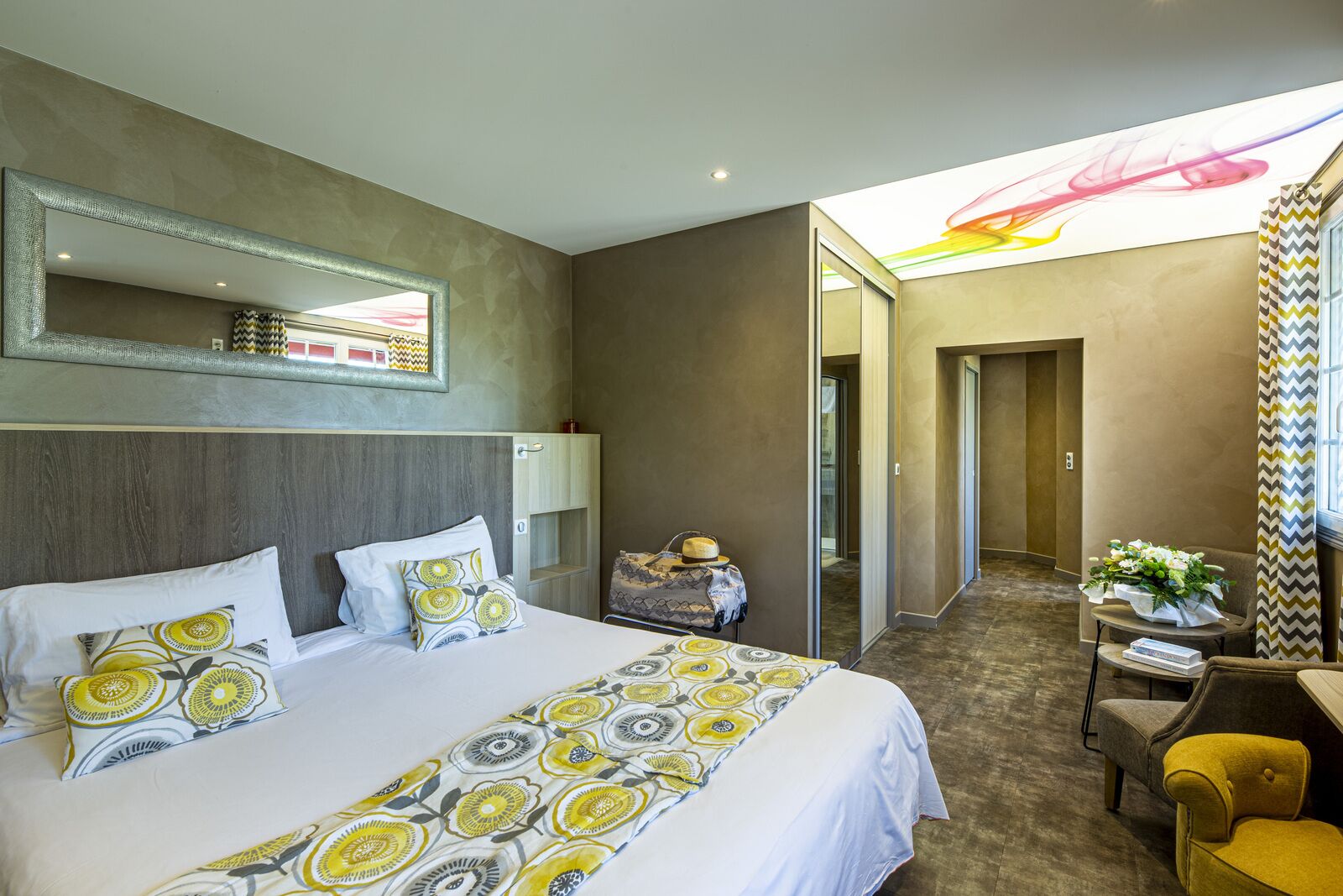 A hotel room with a large bed, a desk, and a chair. There is a mirror on the wall and a window with a view of the sky.