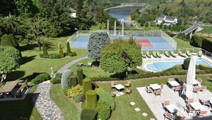 A landscaped outdoor area with a tennis court, seating areas, and a view of a lake and hills. - Hôtel Beau Site (Val-d'Arcomie)