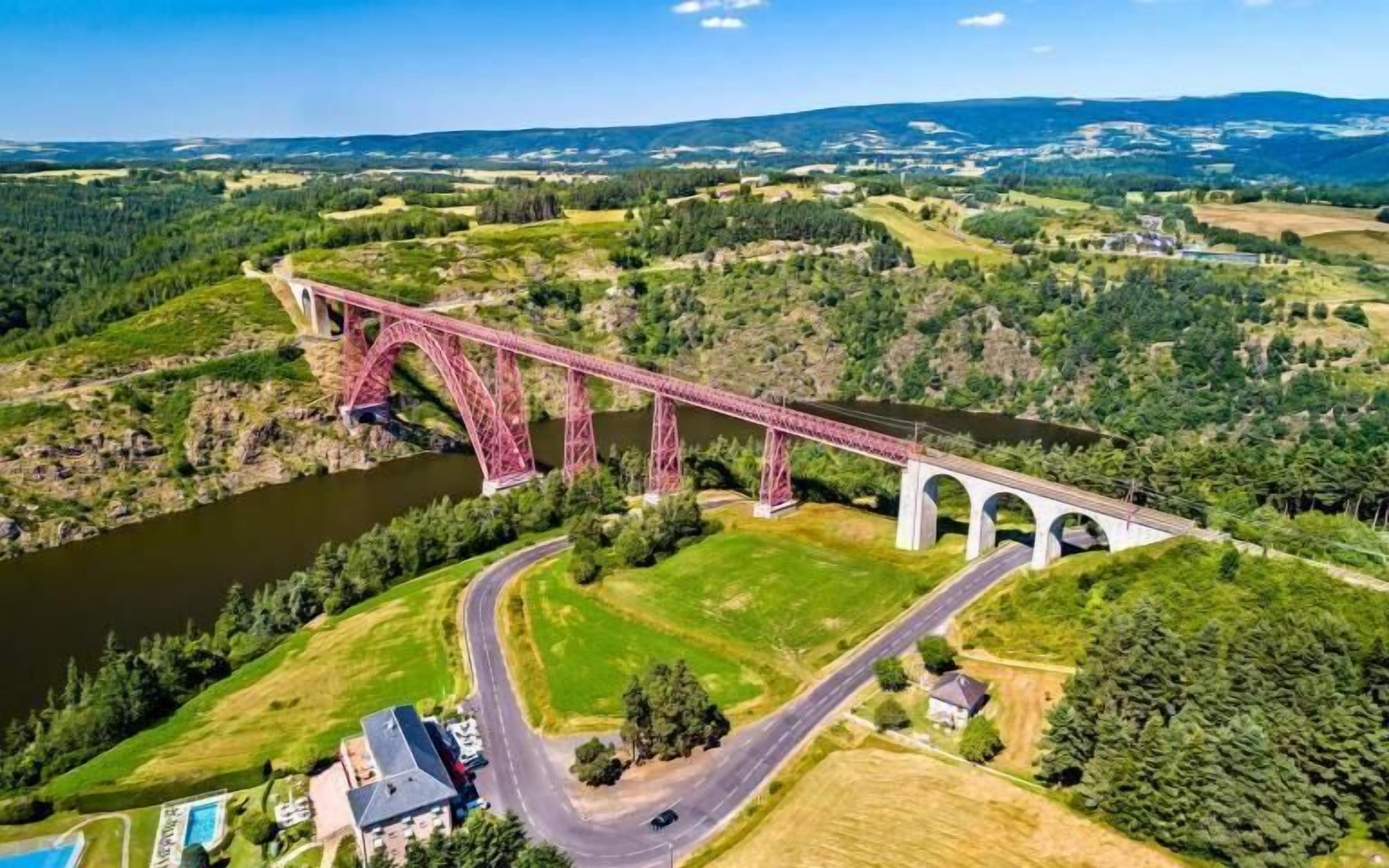 Un ponte rosso sopra un fiume, con una strada che lo costeggia, immerso in campi verdi e alberi.