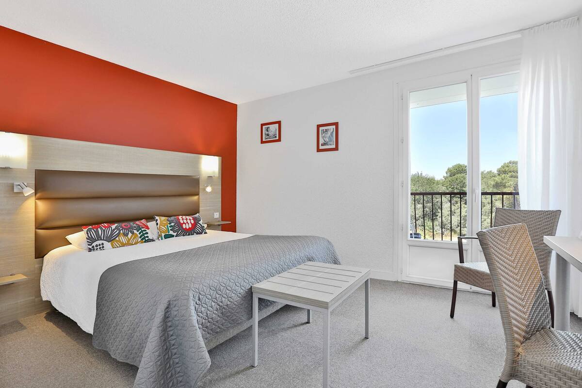 Habitación de hotel con cama, una mesita, una silla, balcón con barandilla y dos cuadros en la pared.