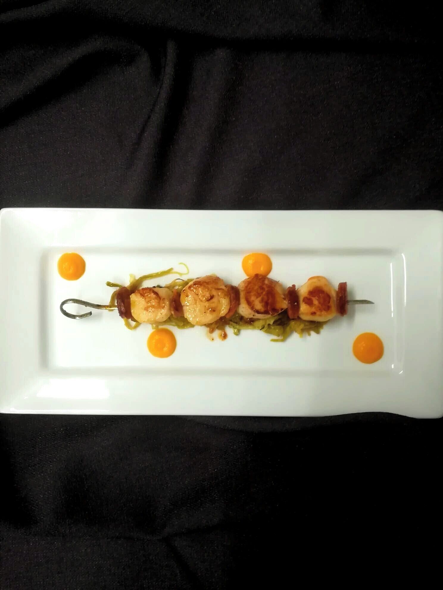 A white rectangular plate with a skewered dish of scallops and vegetables, garnished with small orange dots.