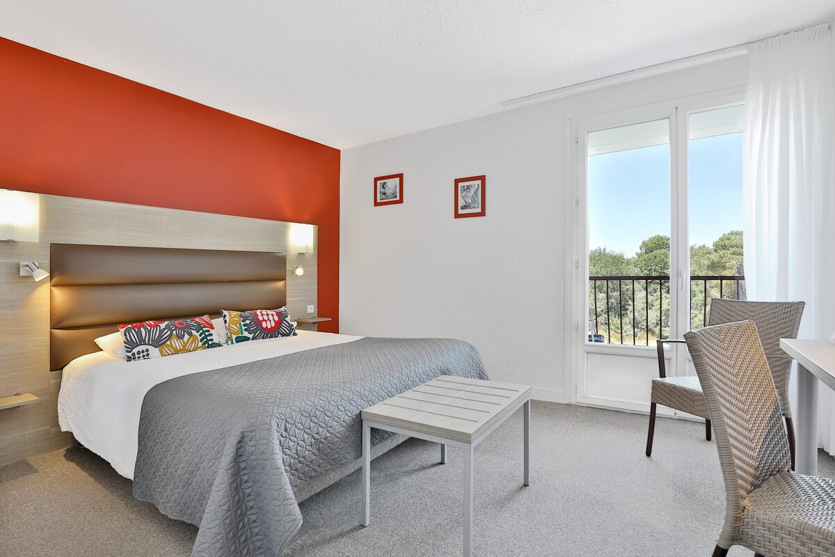 Habitación de hotel con cama, una mesita, una silla, balcón con barandilla y dos cuadros en la pared.
