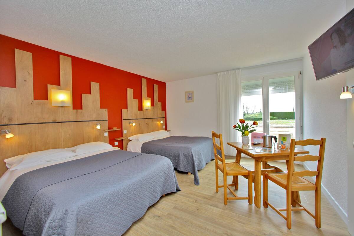 Una habitación de hotel con dos camas, una mesa de madera con sillas, una ventana con cortinas y un televisor montado en la pared.