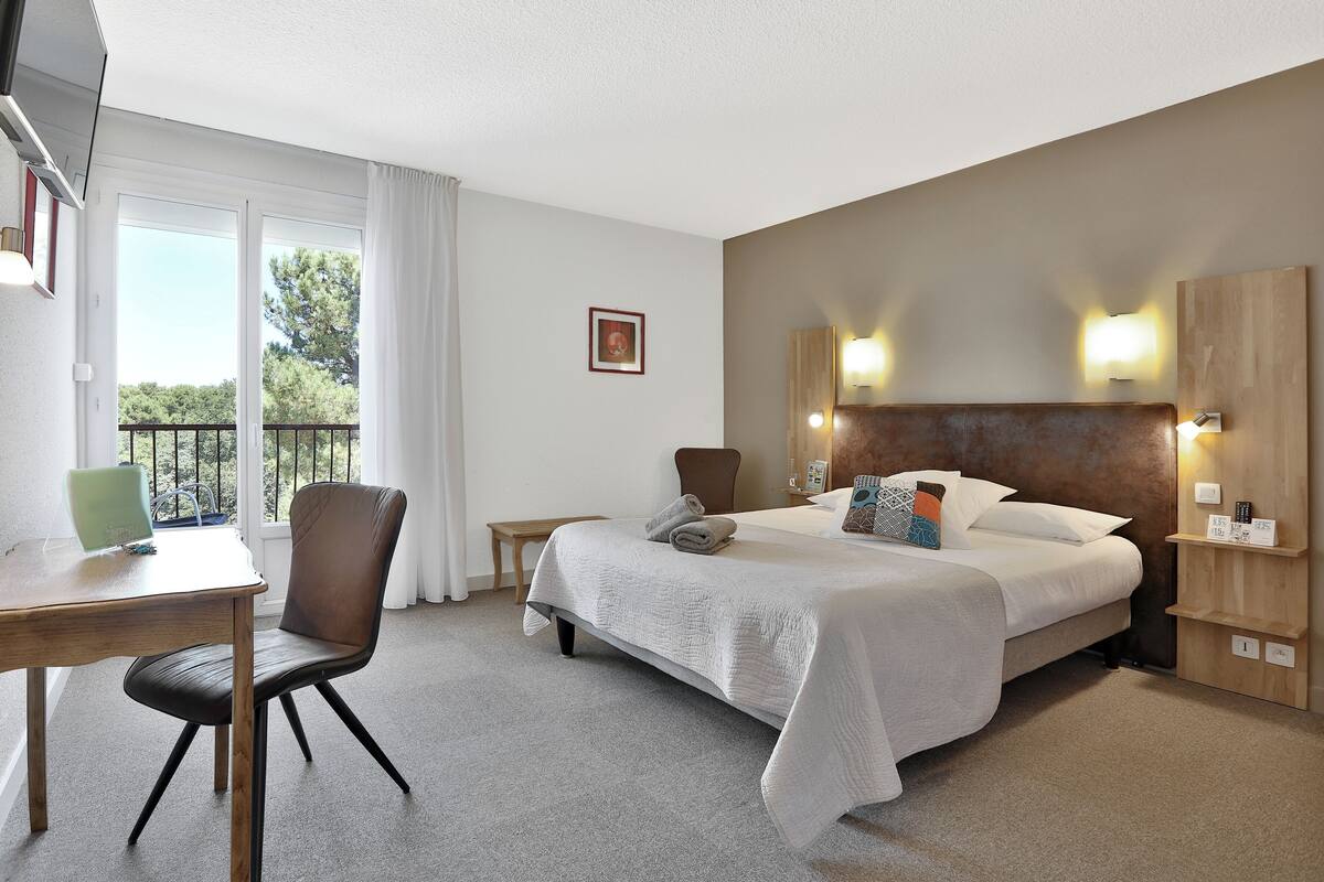 Habitación de hotel con cama, escritorio, silla y balcón con barandilla.
