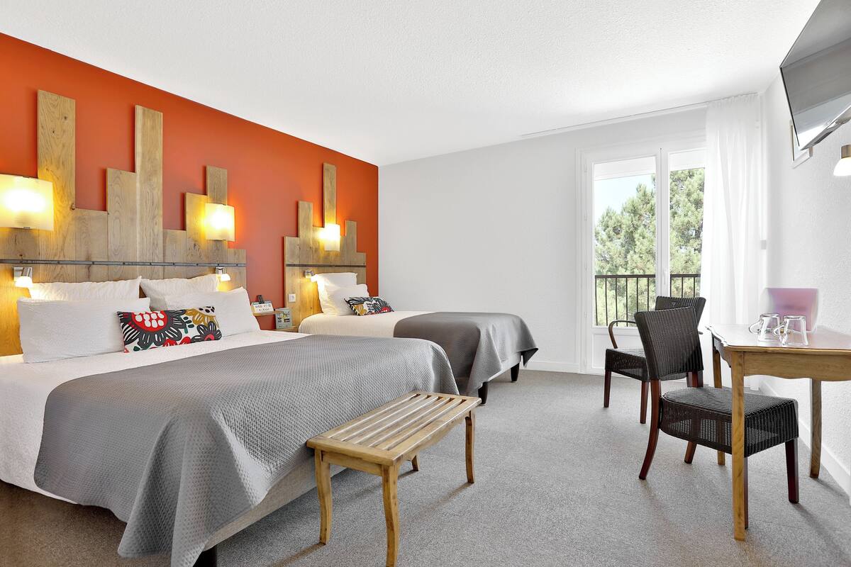 Habitación de hotel con dos camas, un banco de madera, una mesa con sillas y un ventanal con vistas a árboles.