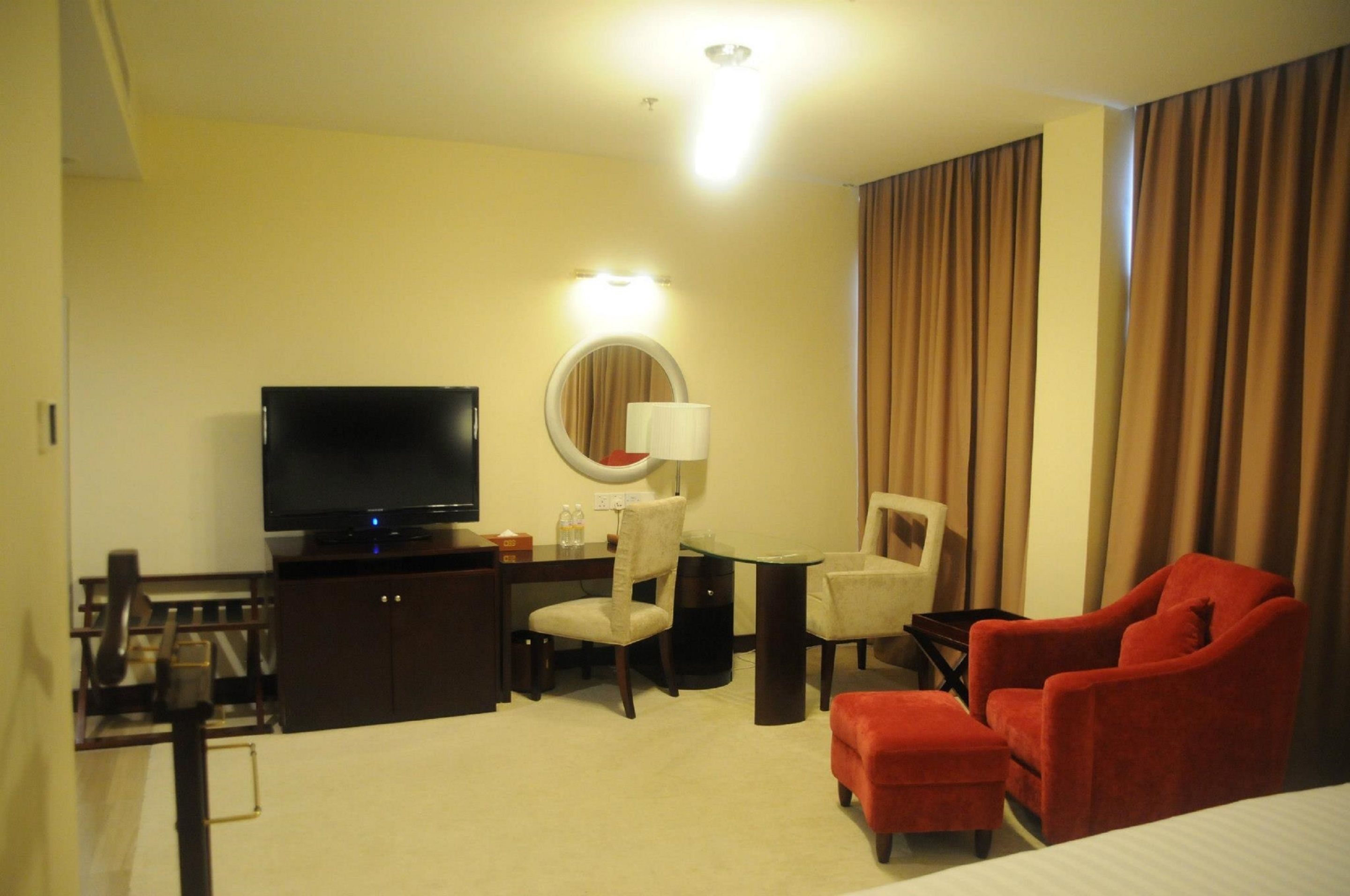 A hotel room with a television, a round mirror, a desk with a lamp, two chairs, and a red armchair.