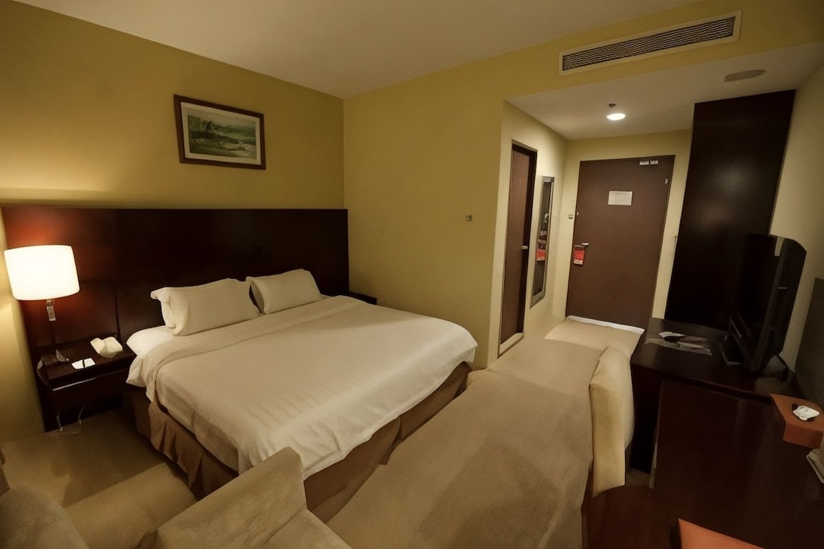 Ett hotellrum med en säng, ett nattduksbord, en lampa, en stol, ett skrivbord, en tv och en dörr.