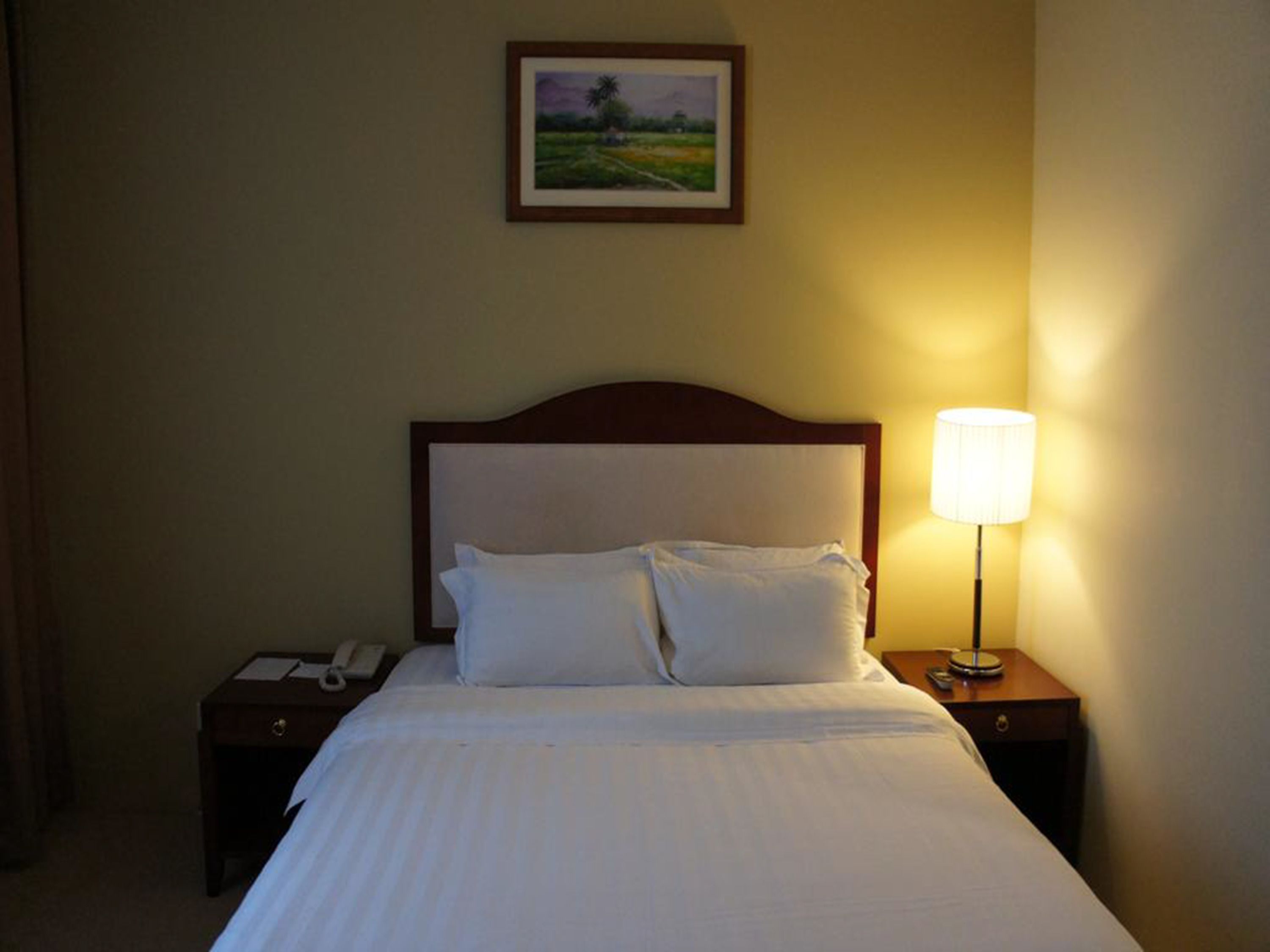 Ein Hotelzimmer mit einem Bett, zwei Nachttischen, einer Lampe, einem gerahmten Bild an der Wand und einem Telefon auf einem der Tische.