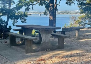 Una mesa de picnic con bancos, cerca del agua y con un puente al fondo.