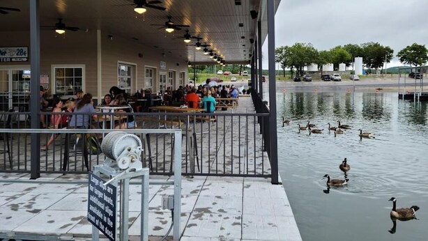 A covered outdoor seating area with tables and chairs, a sign indicating a 28% discount, and ducks swimming in the water. - Port of Kimberling Marina & Resort (Kimberling City)
