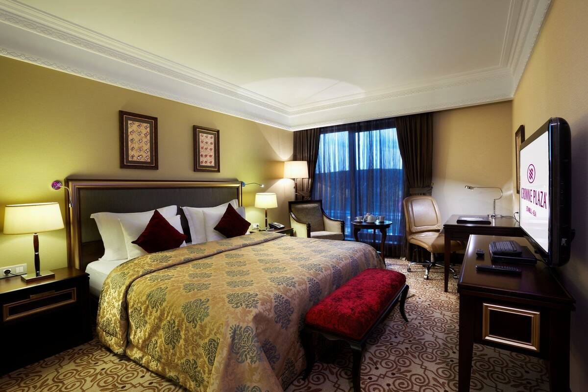 A hotel room with a large bed, a desk with a computer, a television, and a window with curtains.