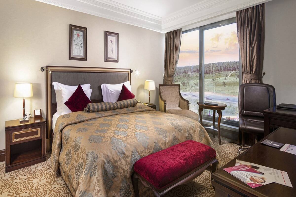 A hotel room with a large bed, two bedside tables, a chair, a desk, and a view of a landscape through a large window.