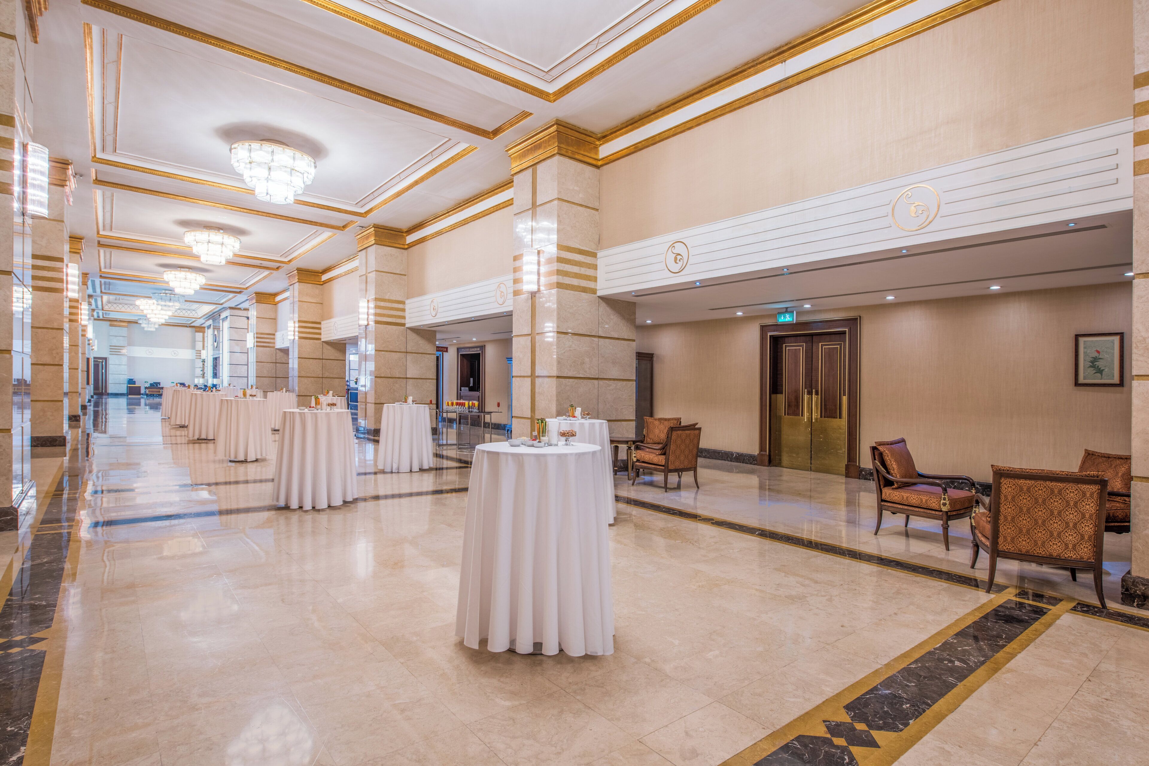 A spacious hotel corridor with multiple round tables set up for dining, a reception area with a counter, and a seating area with chairs.