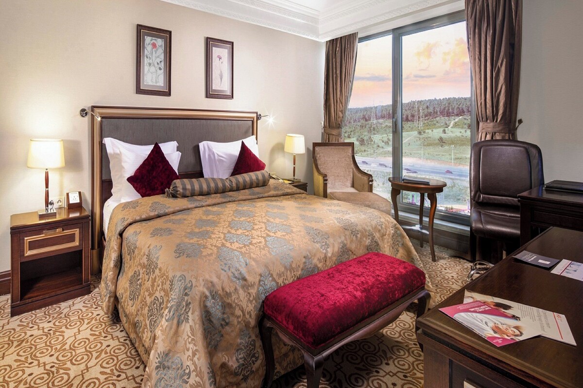 Hotelkamer met een groot bed, twee nachtkastjes, een stoel, een bureau en uitzicht op een landschap door een groot raam.