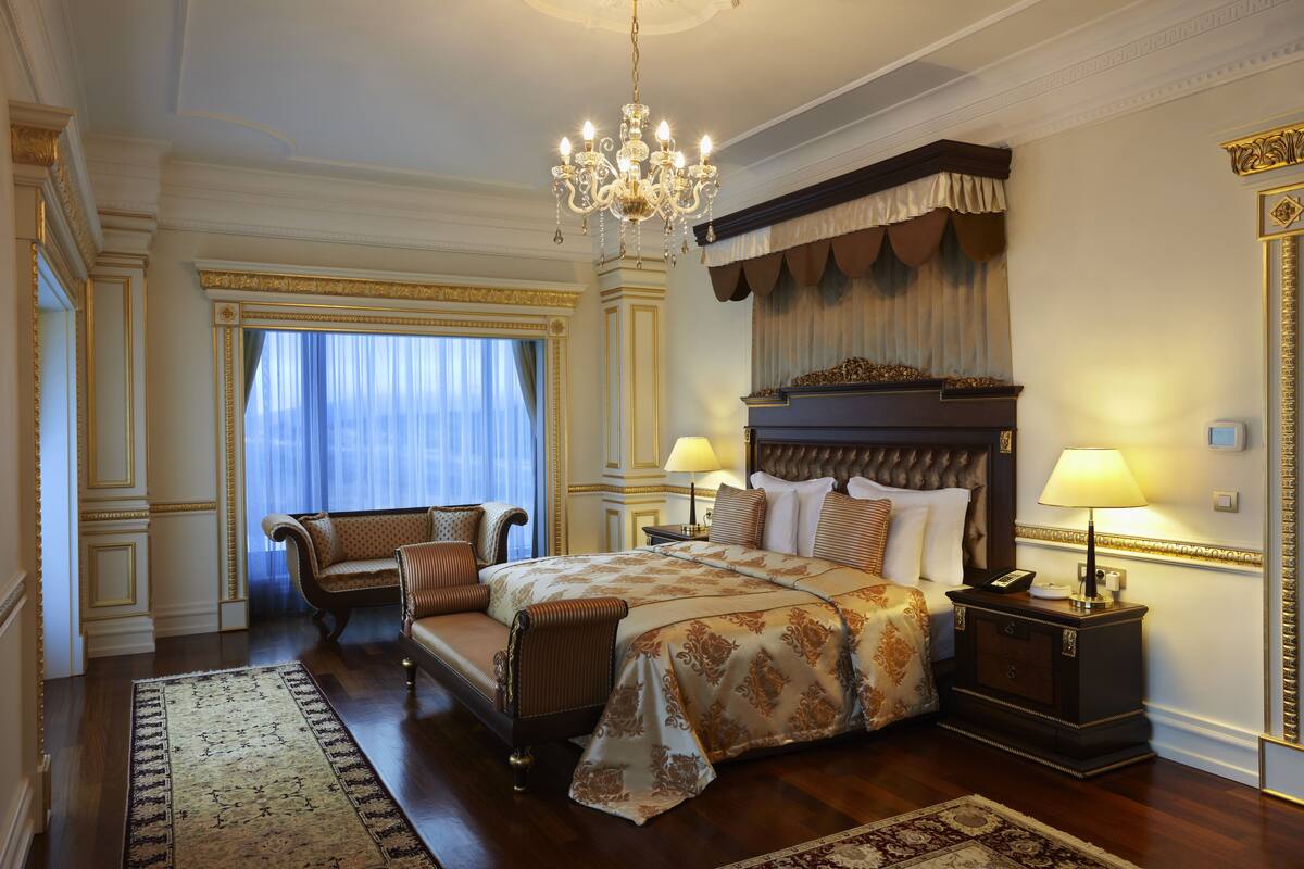 A luxurious bedroom with a large bed, a sitting area with a sofa, a chandelier, and a nightstand with a lamp.