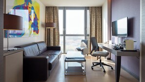 A modern hotel room with a leather sofa, a desk with a computer, a flat-screen TV, and a large window with curtains.