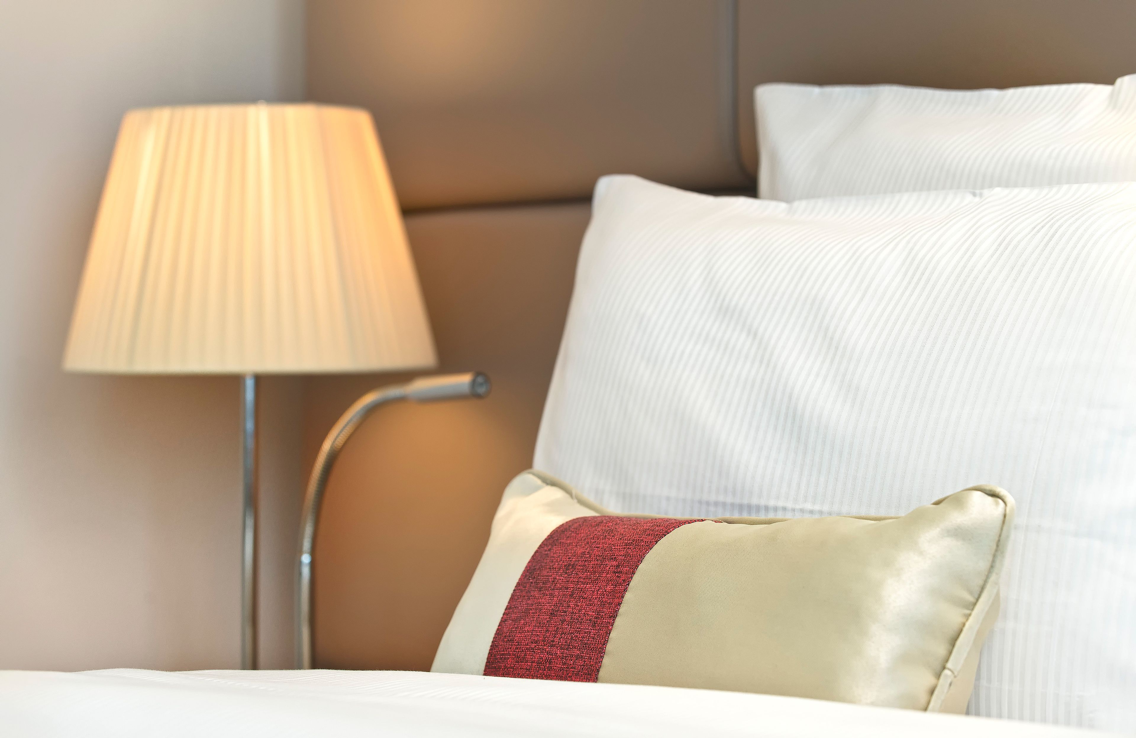 Un lit bien fait, avec une literie blanche et un coussin rouge qui apporte une touche de couleur, une lampe de chevet et une tête de lit.