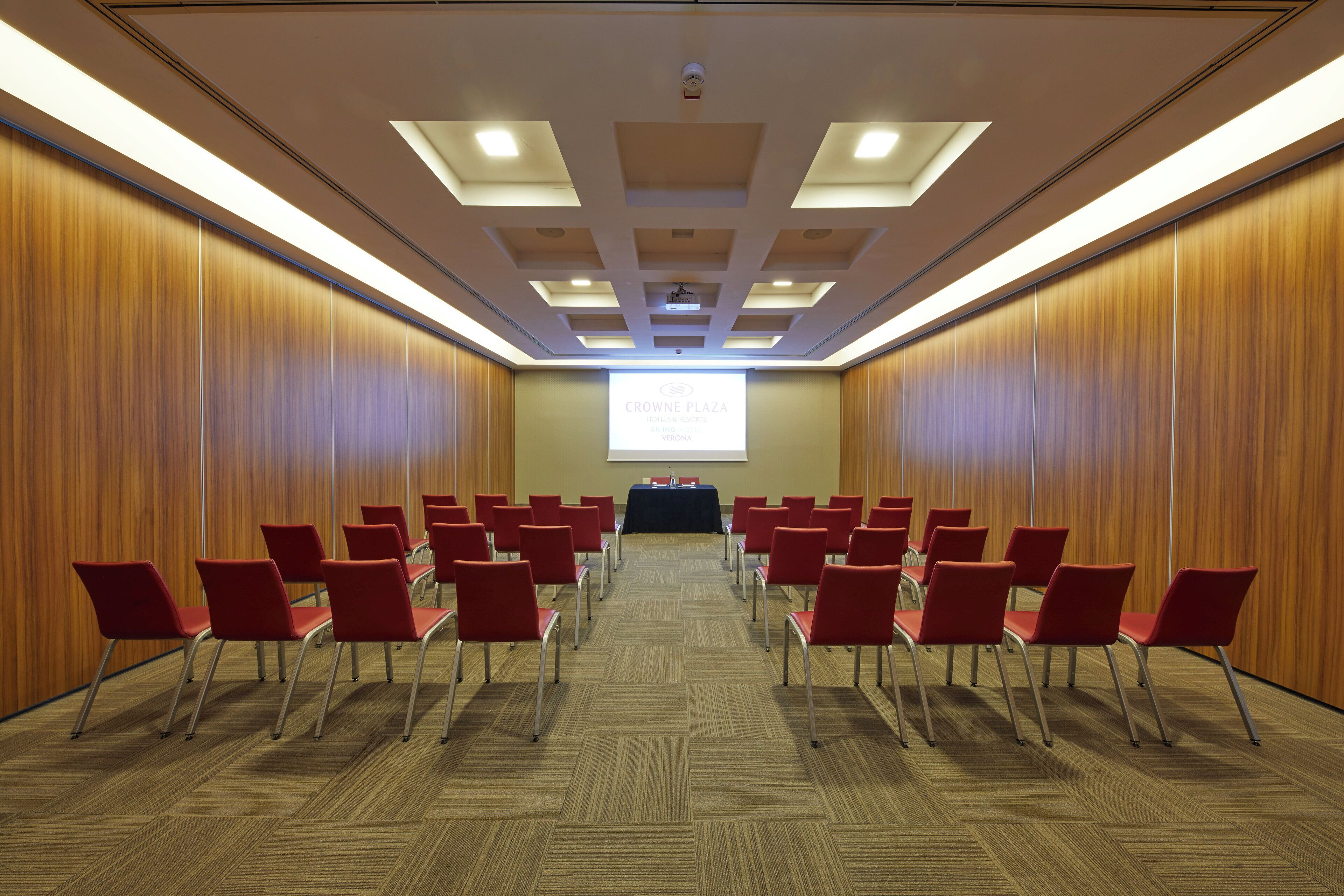 Une salle de conférence avec des rangées de chaises rouges, un pupitre et un écran de présentation.