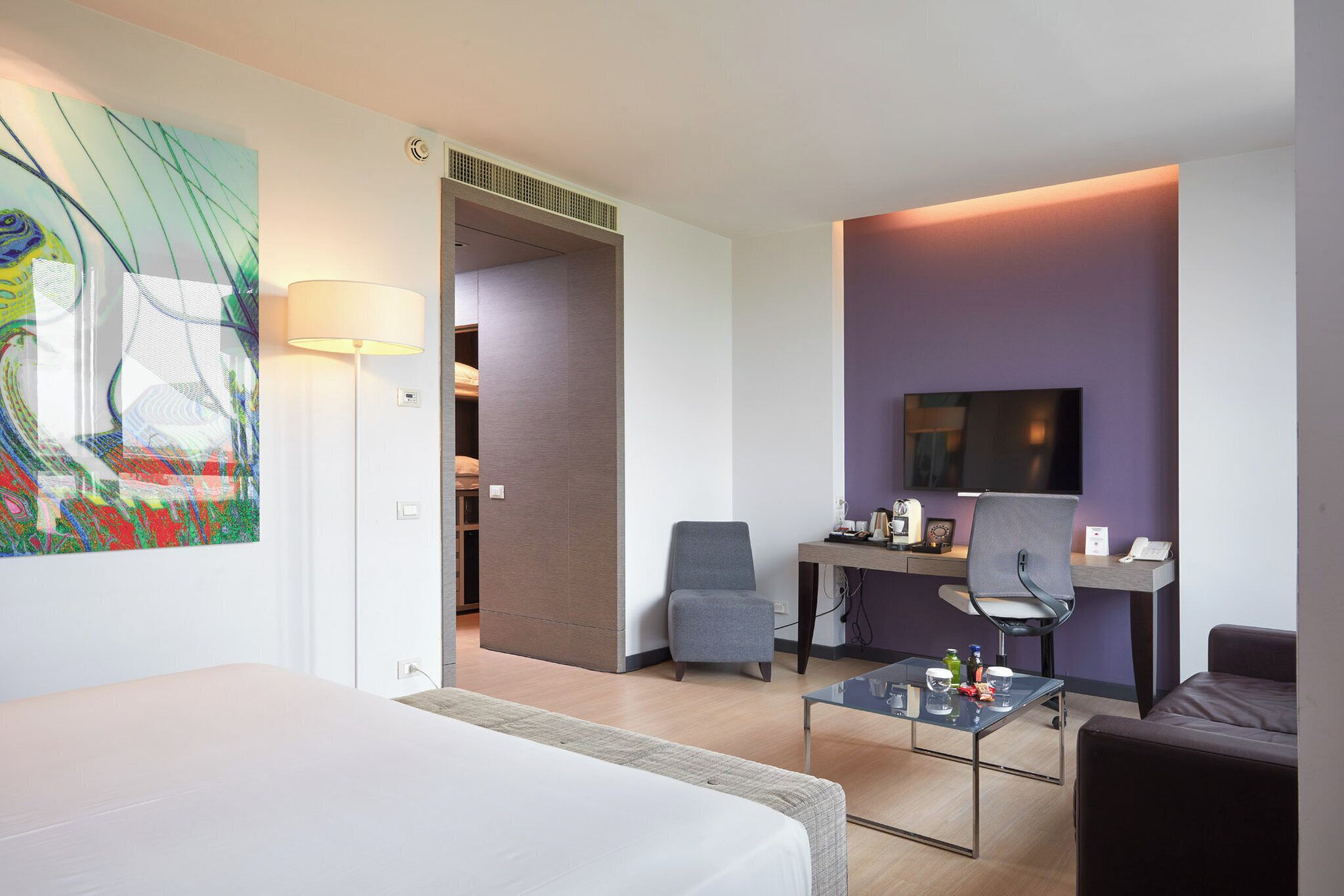 A modern hotel room with a bed, a desk with a chair, a TV, and a painting on the wall.