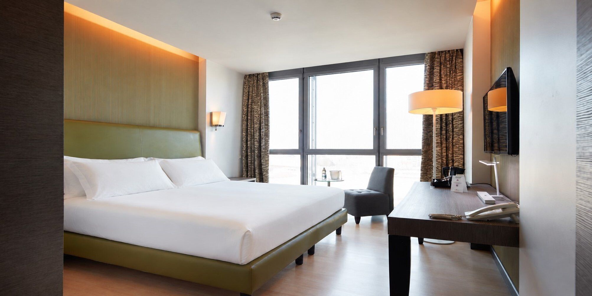 A hotel room with a large bed, a desk, a chair, a TV, and a window with curtains.