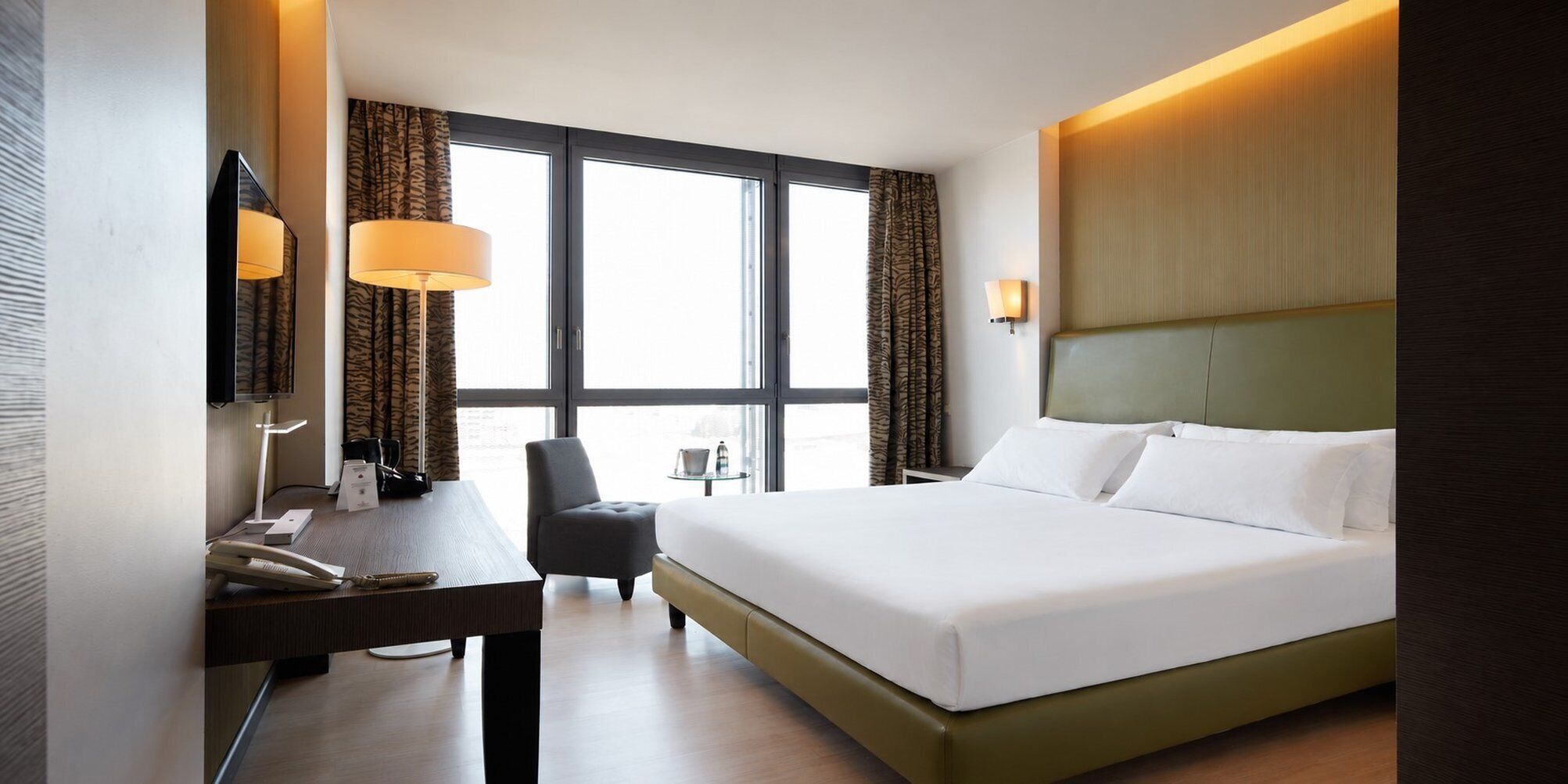 Une chambre d’hôtel avec un grand lit, un bureau avec un téléphone, une chaise et une fenêtre avec des rideaux.