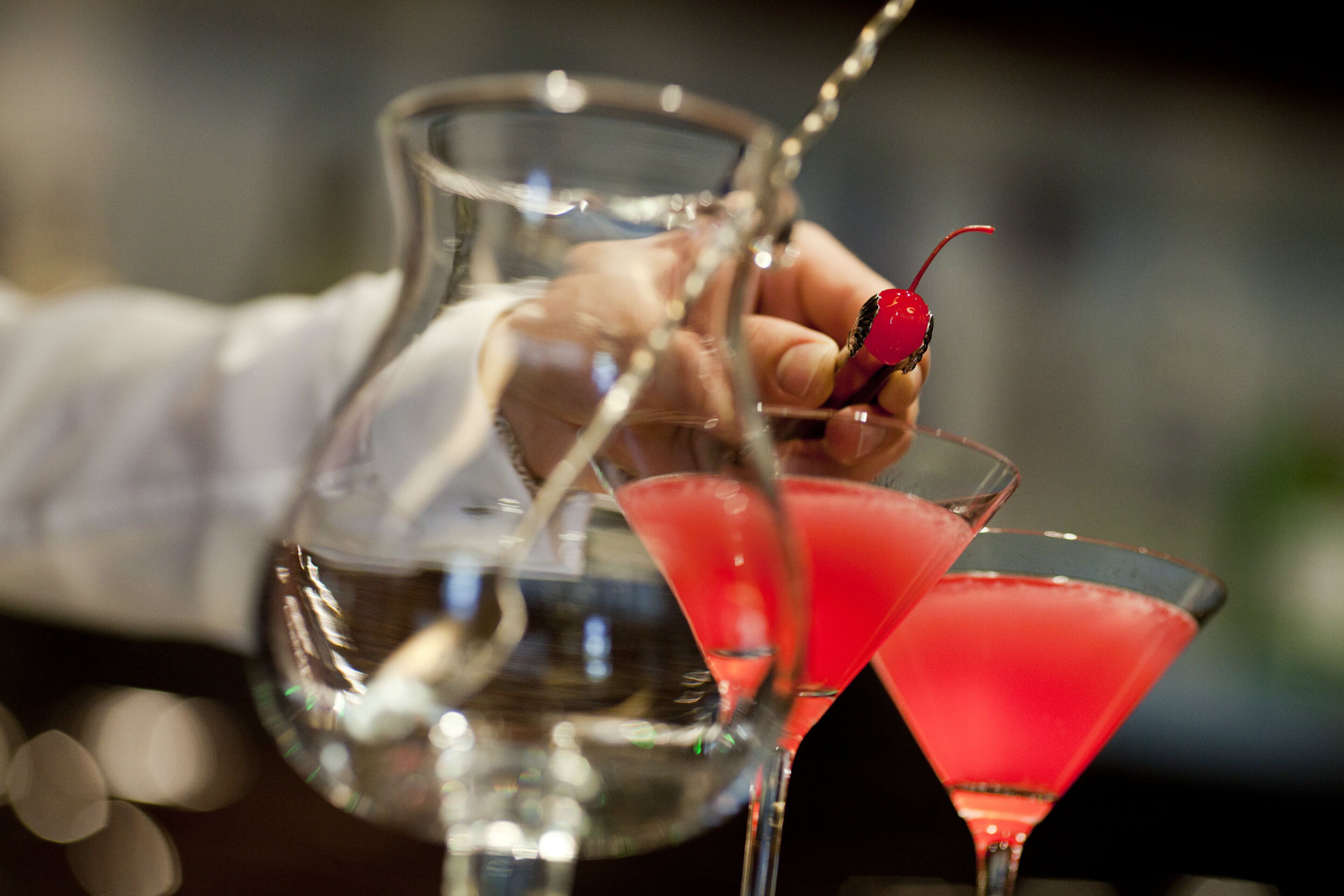 A bartender pouring a drink into a martini glass with a cherry garnish.