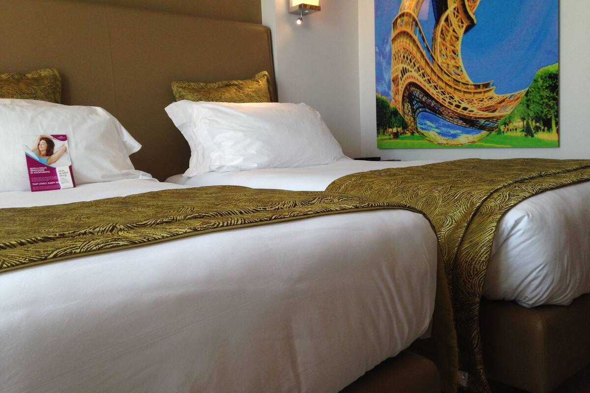 Een hotelkamer met twee bedden, witte lakens en een groot schilderij van de Eiffeltoren.