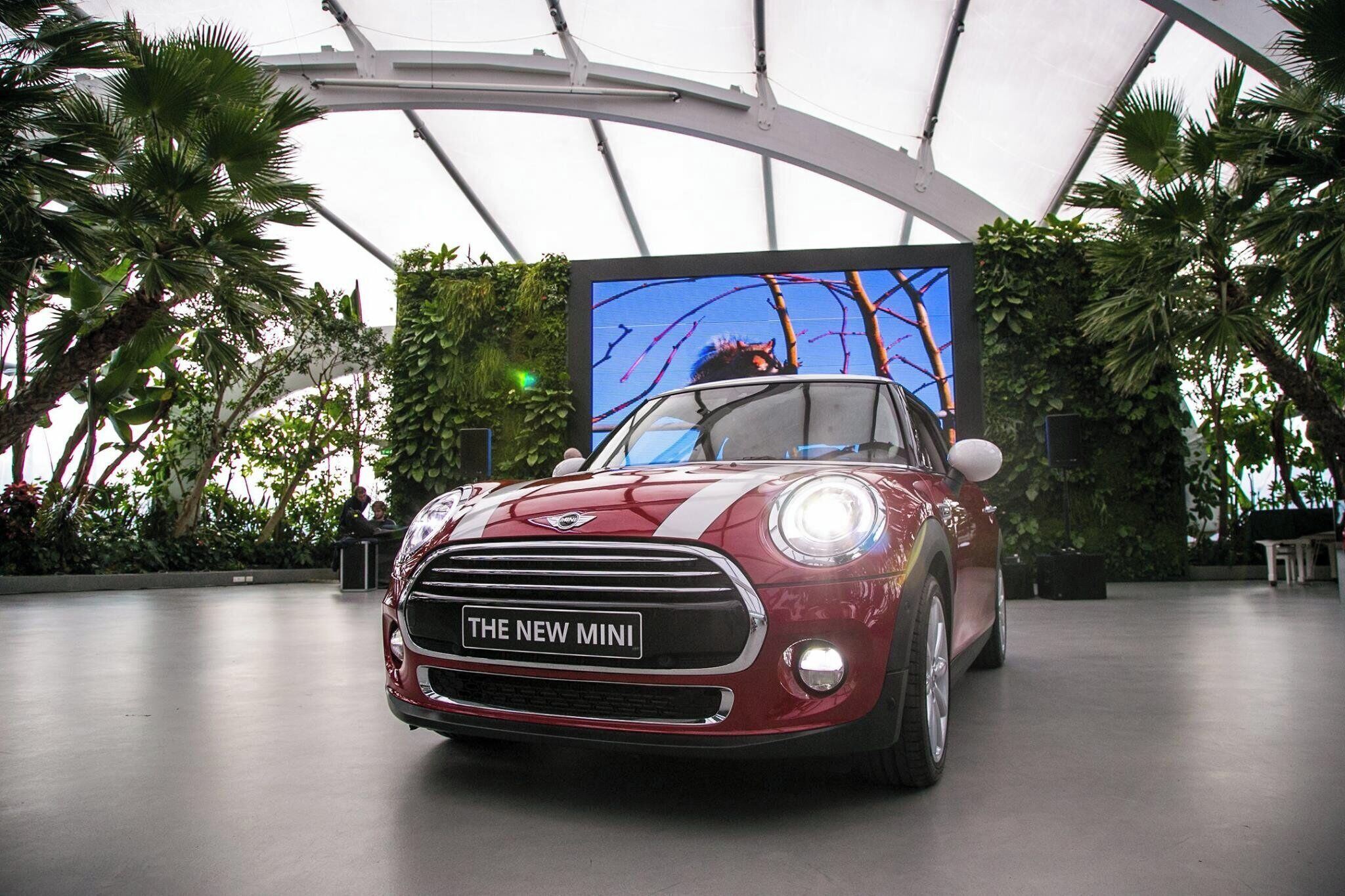 A red Mini car with 'THE NEW MINI' on the front grille, inside a modern building with a curved ceiling and tropical plants.