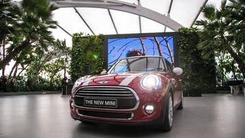 A red Mini car with 'THE NEW MINI' on the front grille, inside a modern building with a curved ceiling and tropical plants.