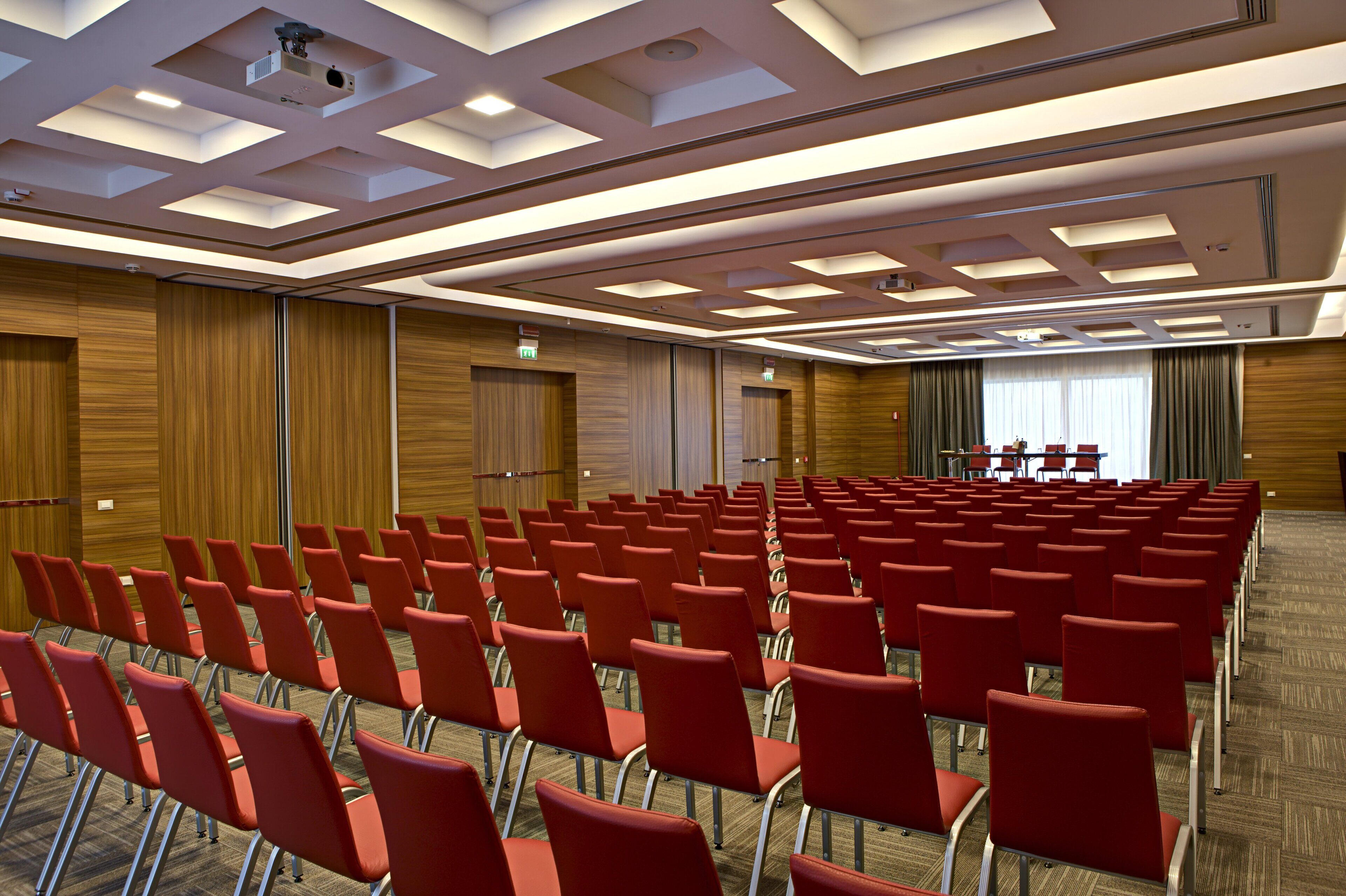 A conference room with rows of red chairs, wooden panel walls, and a ceiling with recessed lighting.