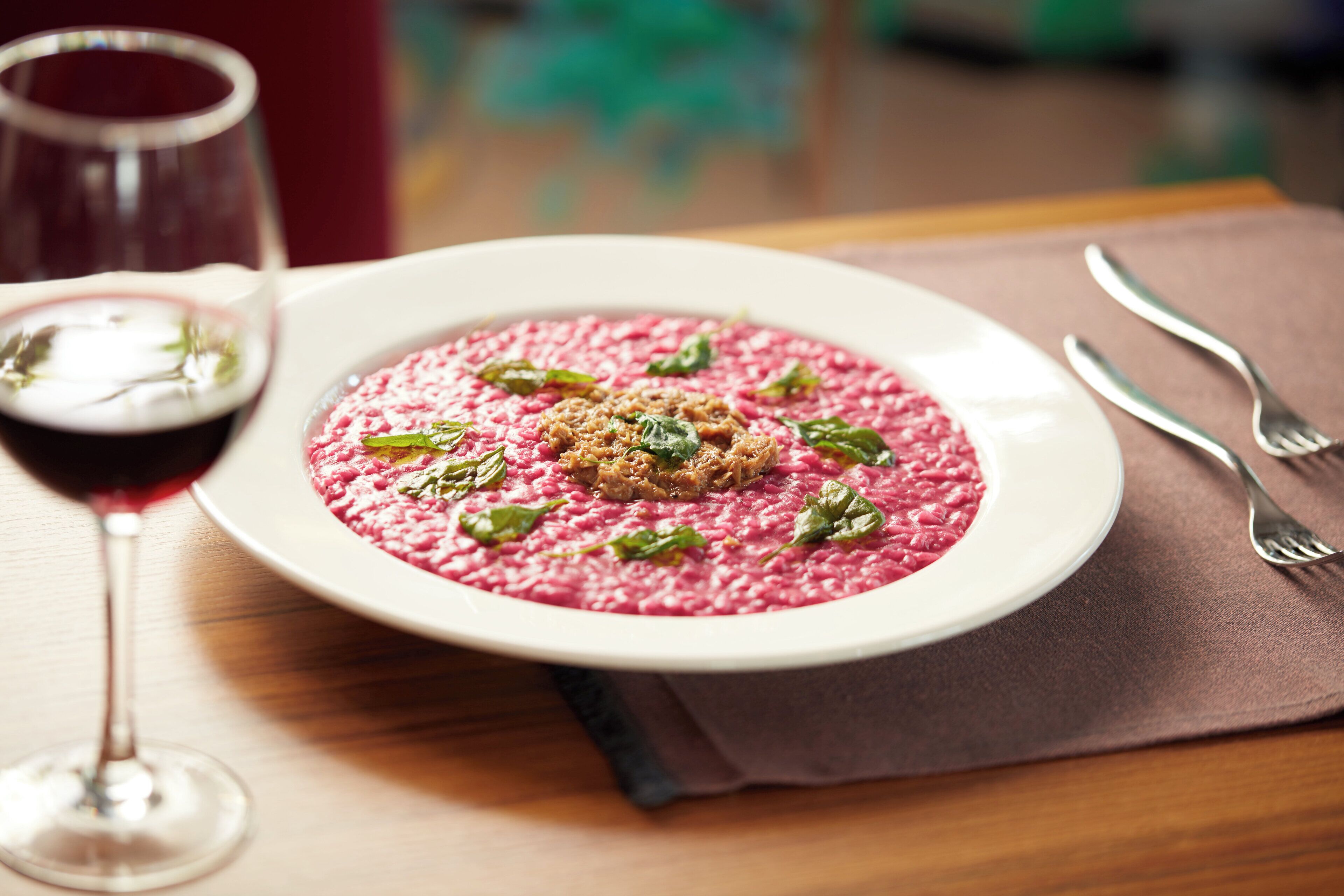 A plate of beetroot risotto garnished with herbs, accompanied by a glass of red wine and cutlery on a wooden table.