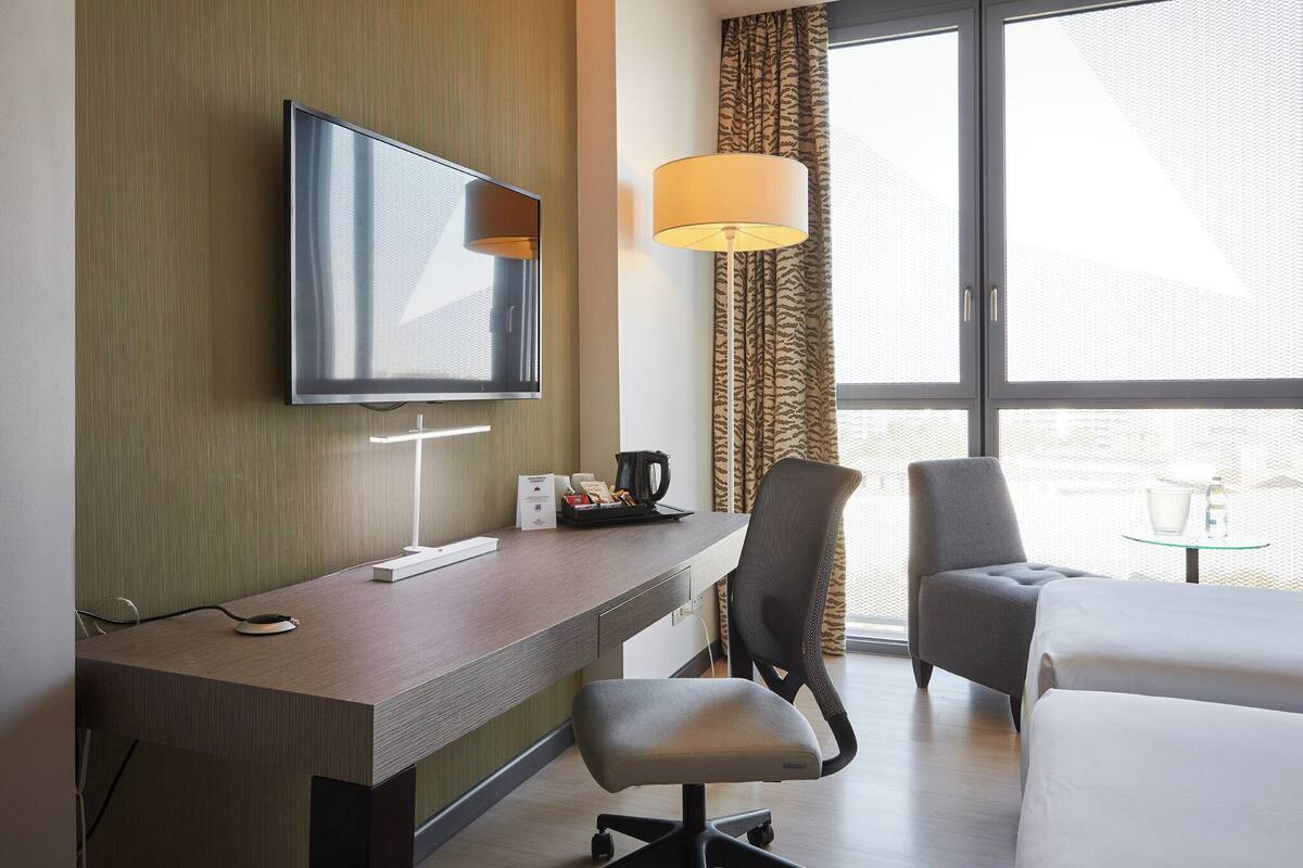 Hotelkamer met een bureau, stoel, bed, televisie, lamp en raam met gordijnen.