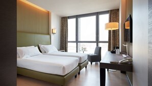 A hotel room with two beds, a desk, a chair, a lamp, a telephone, and a large window with curtains.