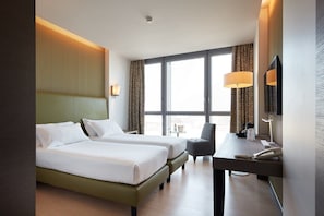 Une chambre d’hôtel avec deux lits, un bureau, une chaise, une lampe, un téléphone et une grande fenêtre avec des rideaux.