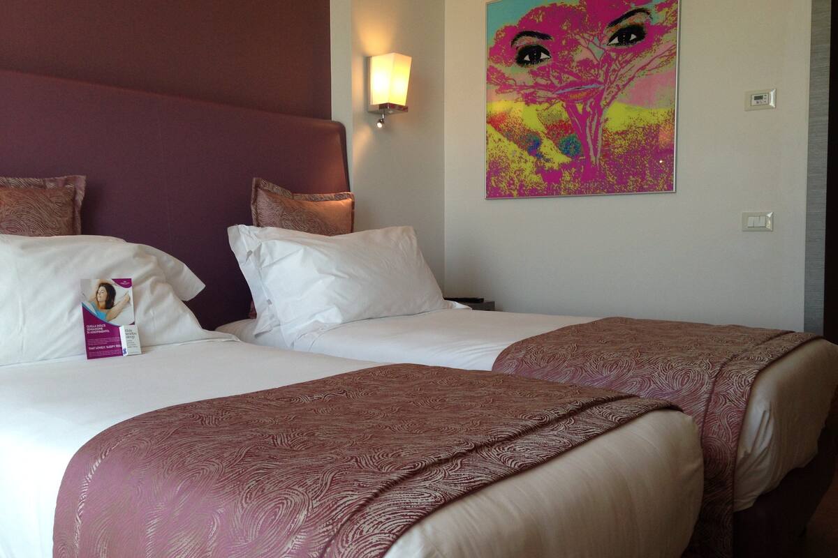 Een hotelkamer met twee bedden, een hoofdeinde, een wandlamp en een kleurrijk abstract schilderij.