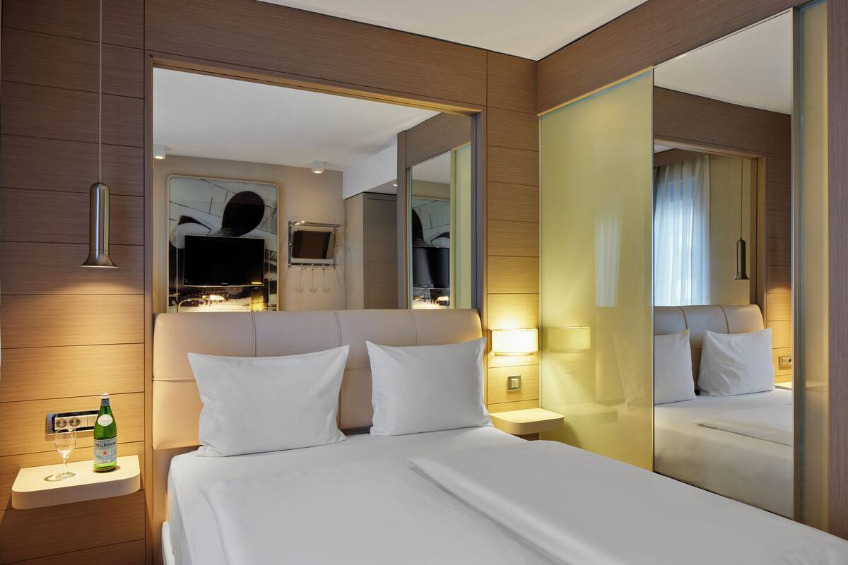 Quarto de hotel com uma cama grande, uma mesa de cabeceira com uma garrafa de água, uma televisão de tela plana e um espelho refletindo o quarto.